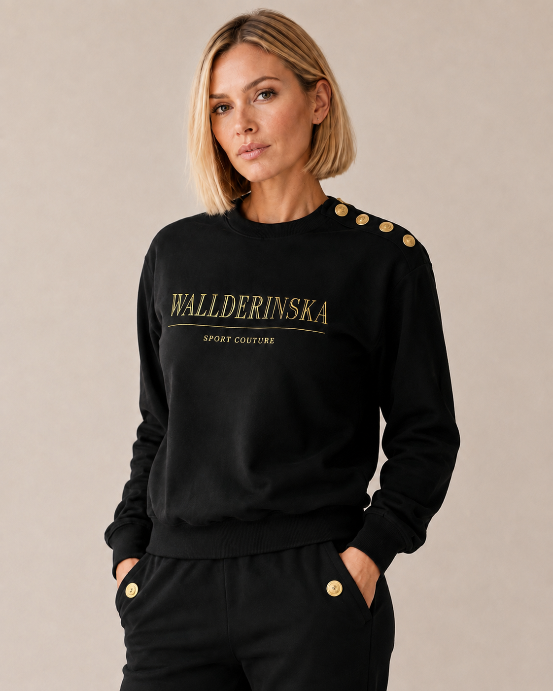 Modellen bär Edge Gold Button Sweater i svart med guldtext på bröstet och guldknappar på ena axeln, till de svarta byxorna med guldknapp vid fickan.