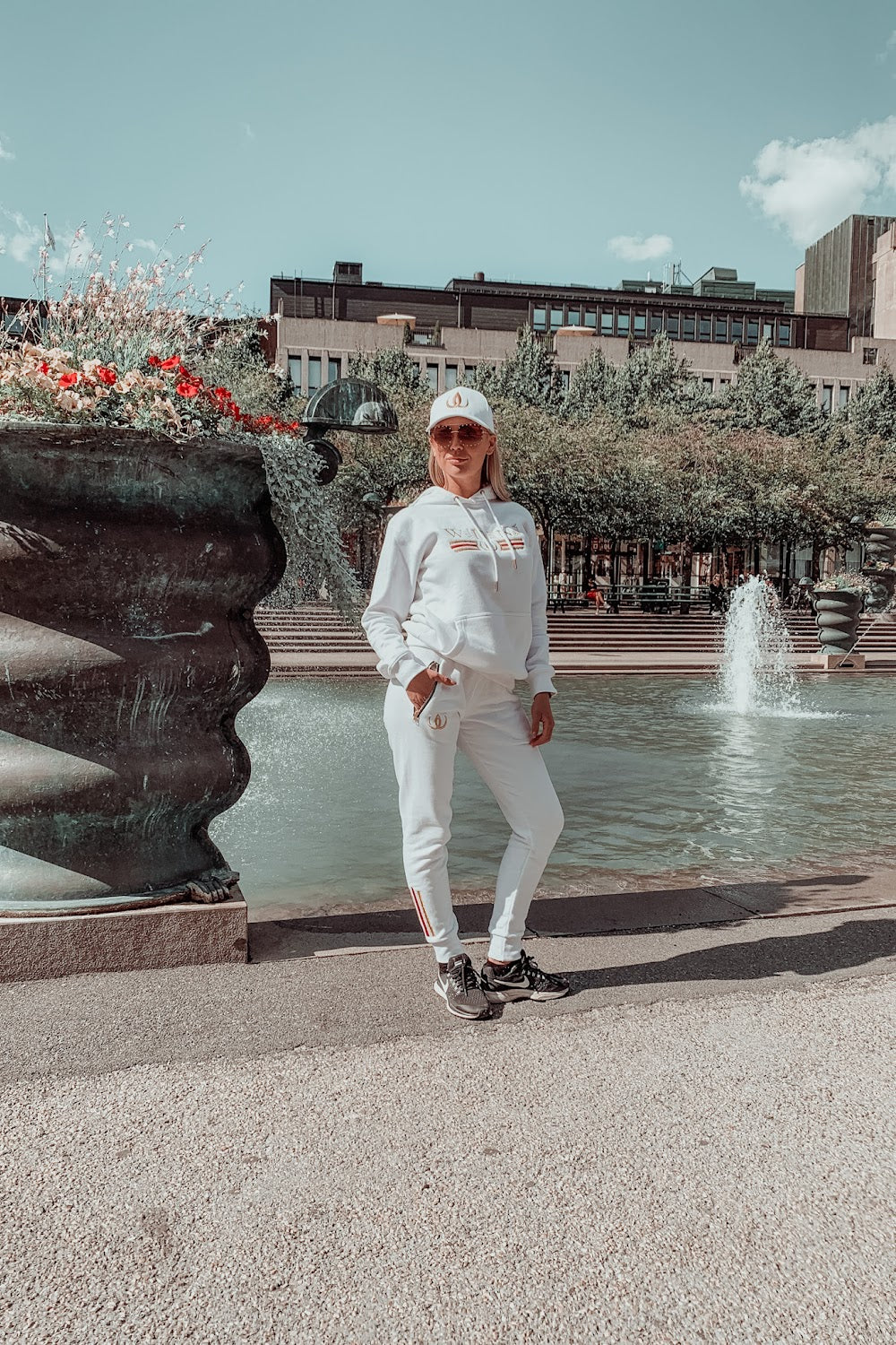Framför fontänen står modellen i vit huvtröja, vita joggers och vit keps.