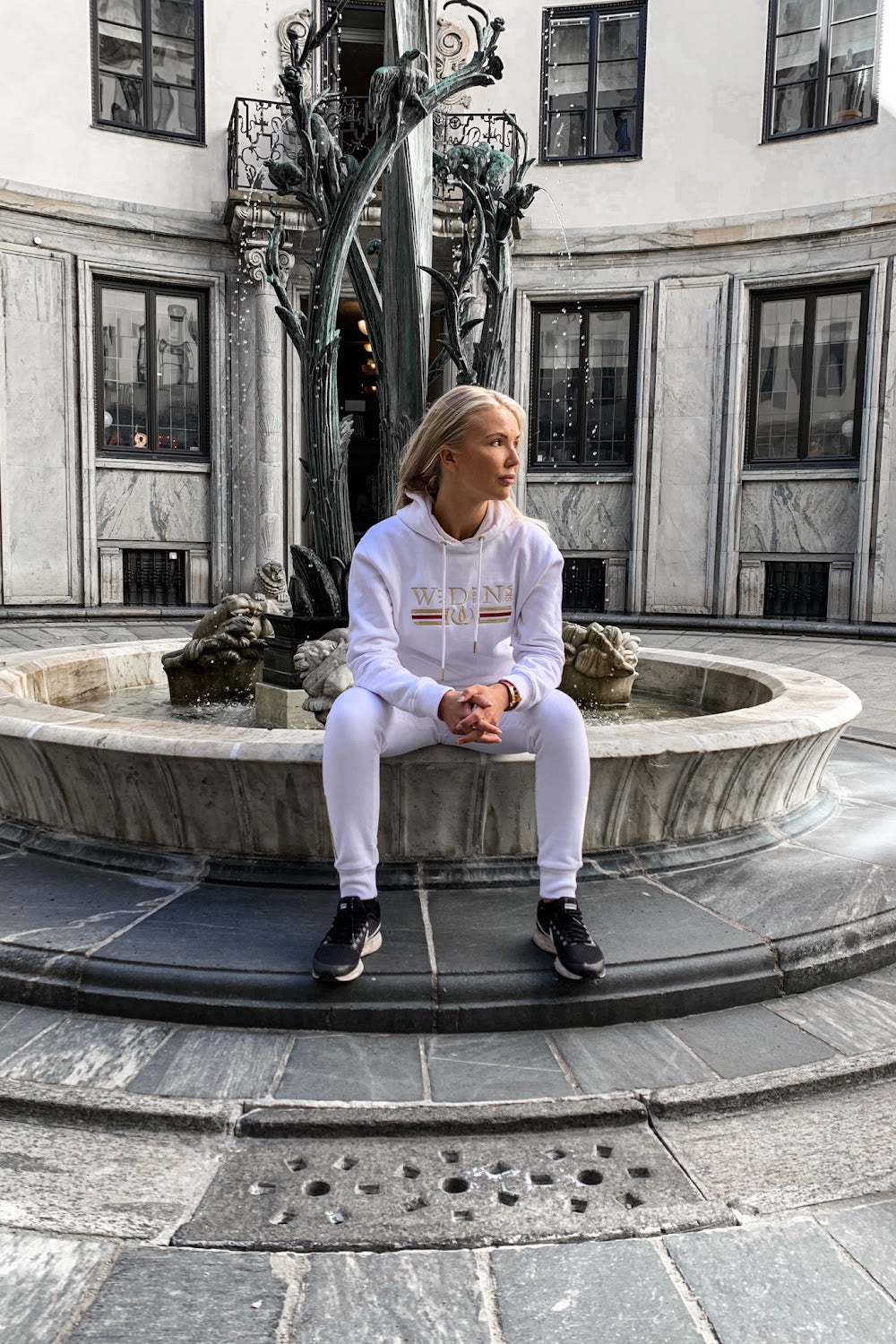 På fontänkanten sitter modellen i den vita huvtröjan, med en staty i bakgrunden.