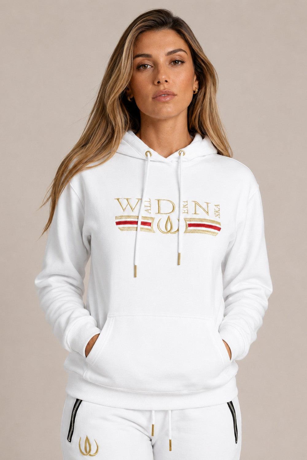 Vit Deluxe Hoodie med guldbroderi och rött streck över bröstet, buren till vita Deluxe Joggers med guldlogga.