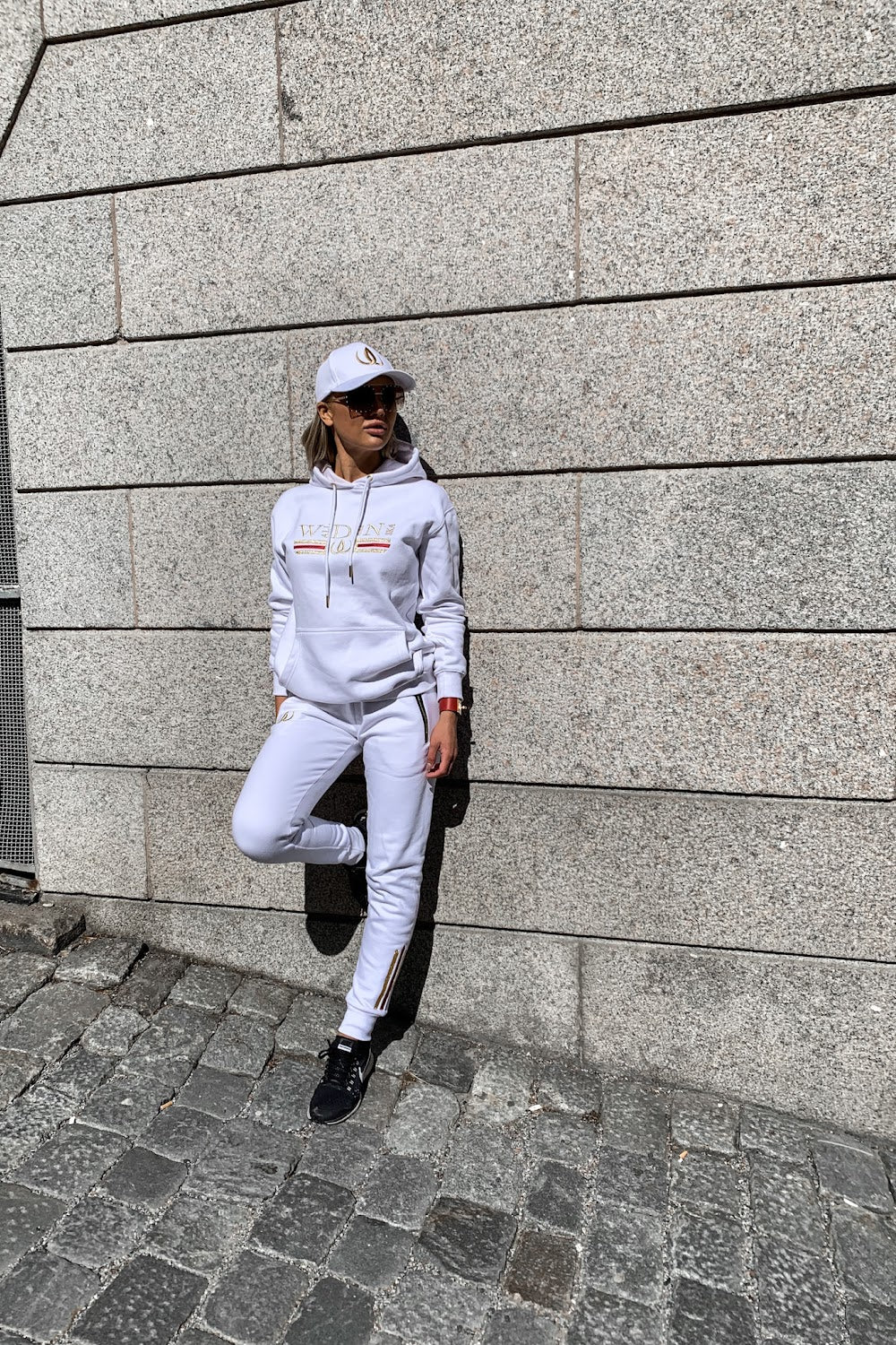 Mot stenväggen lutar modellen sig med ena foten uppdragen, i vit huvtröja och vita joggers.