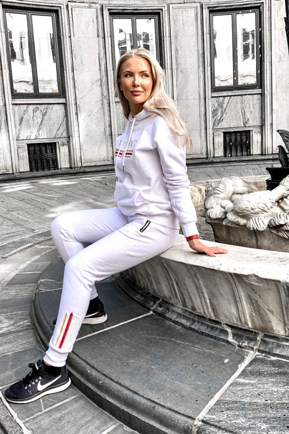 Modellen sitter vid en fontän på stan i den vita huvtröjan och vita joggers med guldrand.