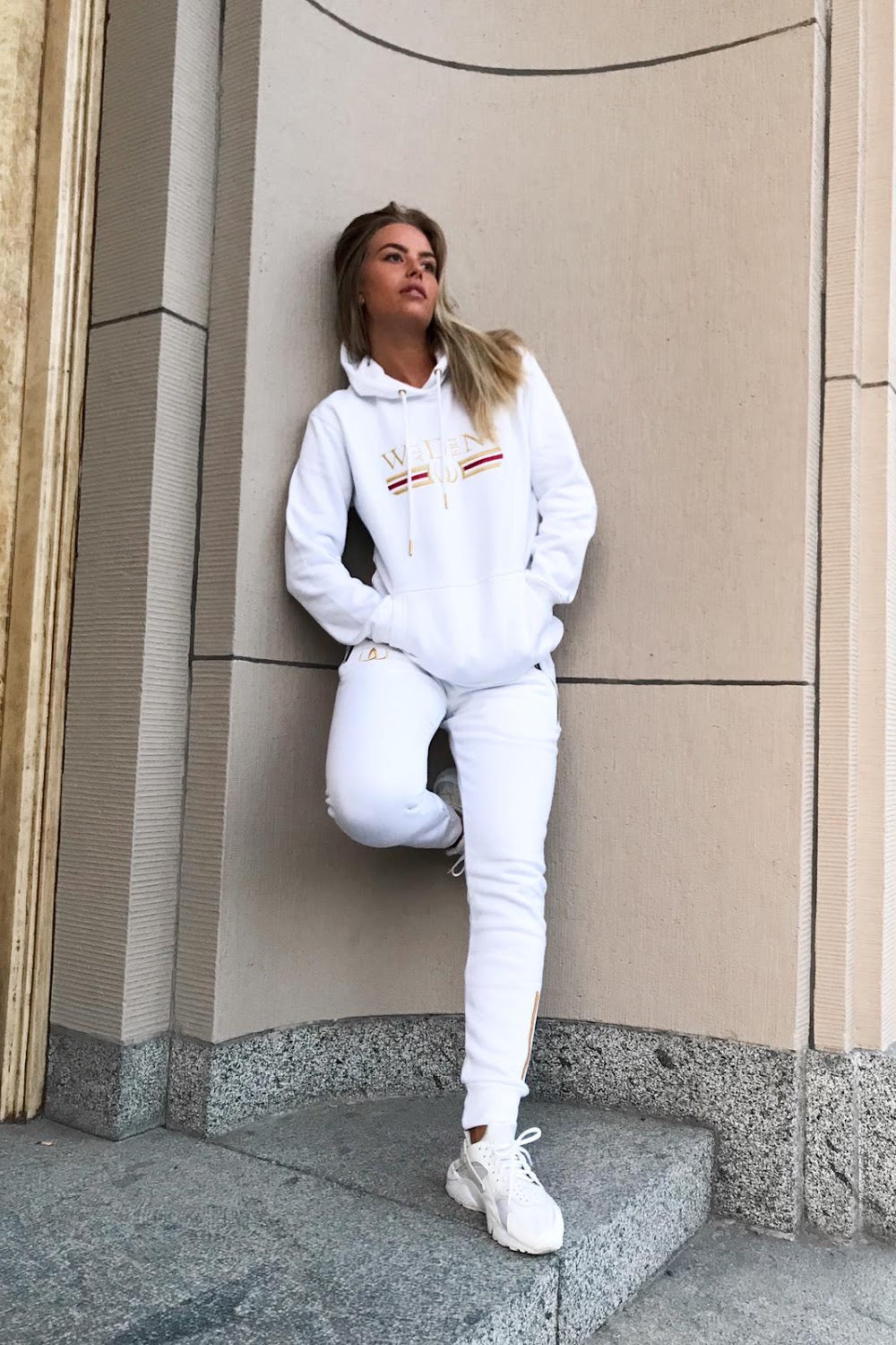 Mot husväggen står modellen i vit huvtröja och vita joggers, till vita sneakers.