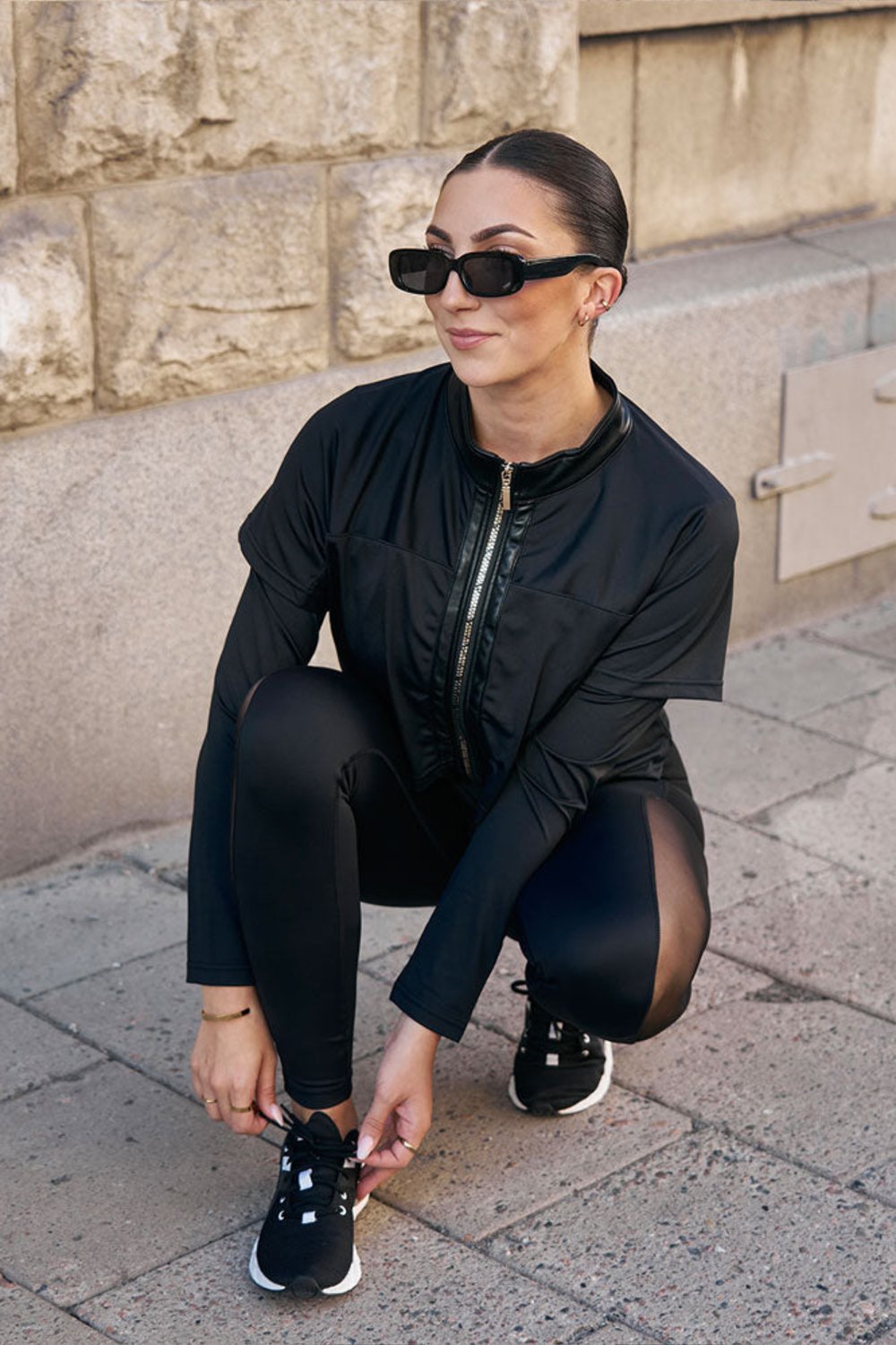 Vid trottoaren knyter modellen skon på huk, i den svarta jackan och svarta tights.